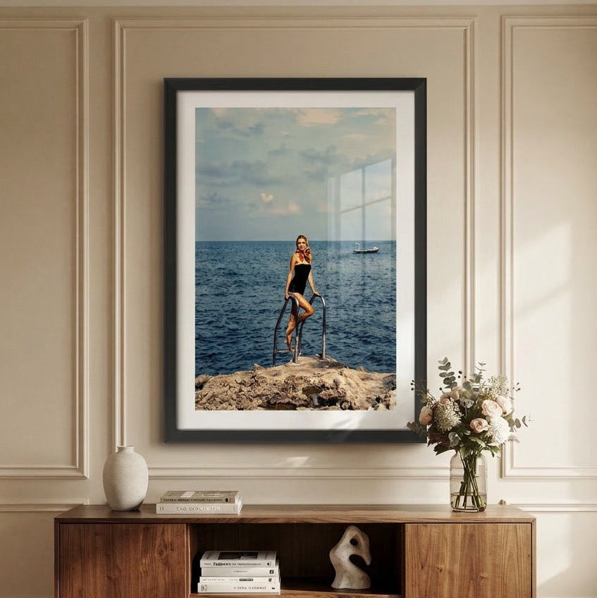 A framed vertical art print depicting a woman standing on a rocky cliff by the sea, displayed on a beige paneled wall above a wooden console table with decorative objects.