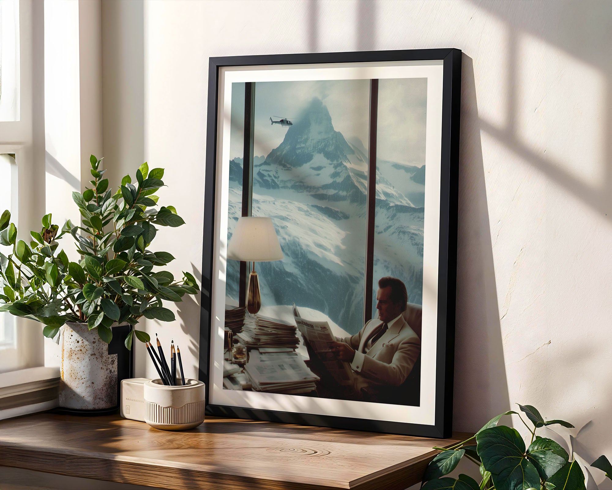 Framed artwork of a man reading a newspaper with mountains in the background, placed on a wooden surface with plants.