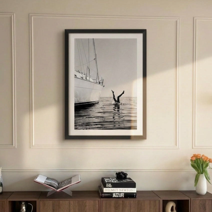 A framed black and white nautical art print featuring a sailboat and a person in the water, displayed on a cream paneled wall above a wooden console cabinet.