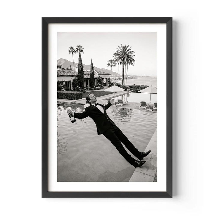 Framed black and white photo of a person in a tuxedo by a pool with palm trees and mountains in the background.