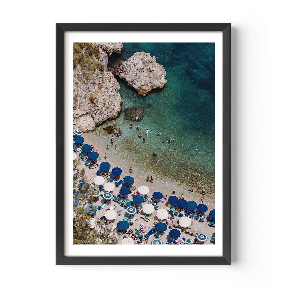 Framed artwork of a beach scene with blue umbrellas and people near rocky cliffs.