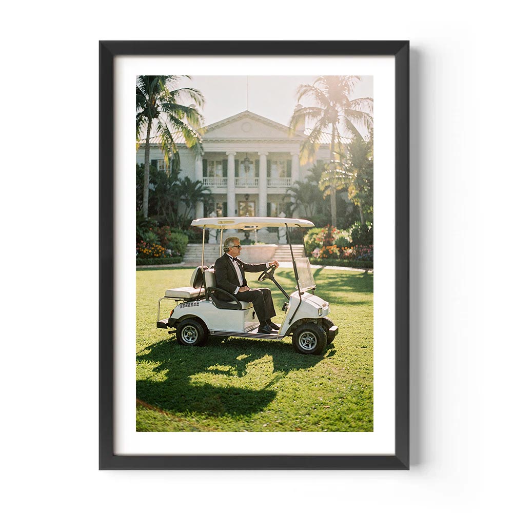 Framed photograph of a person driving a golf cart on a grassy area with palm trees and a building in the background.