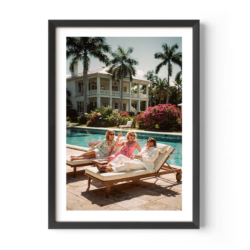 Framed photo of three women lounging by a pool with a mansion and palm trees in the background.