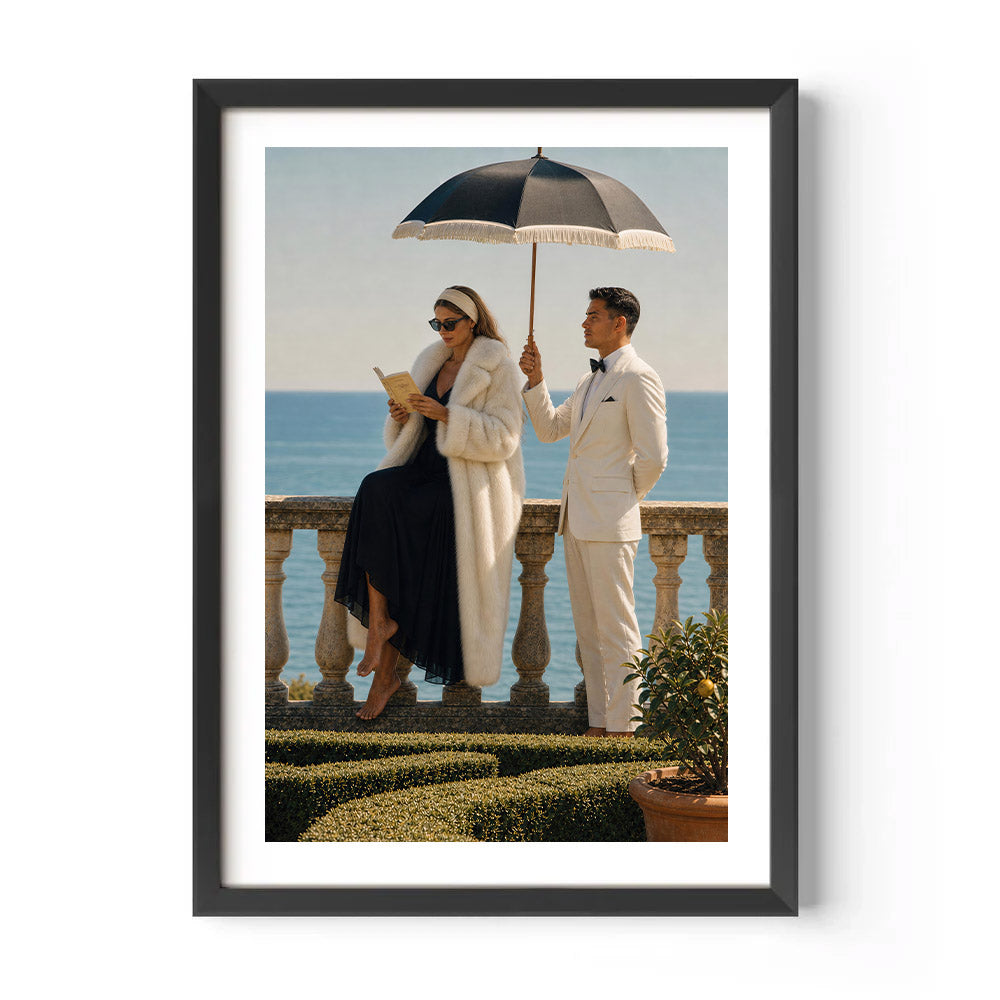 A framed art print featuring a vintage-style photograph of a man and woman on a terrace overlooking the sea. The woman wears a long white fur coat and reads a book under a black parasol, while the man in a white suit stands beside her.