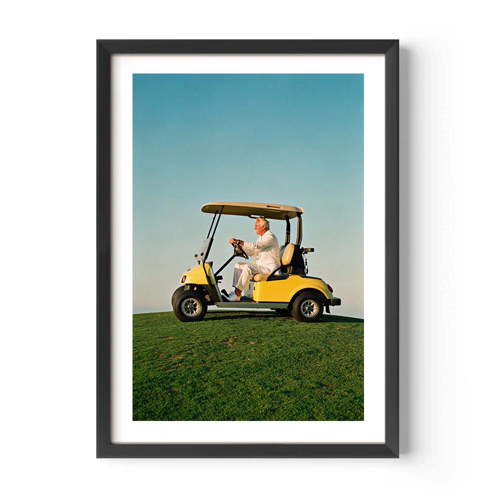 Framed artwork of a man driving a yellow golf cart on a green field with a blue sky.