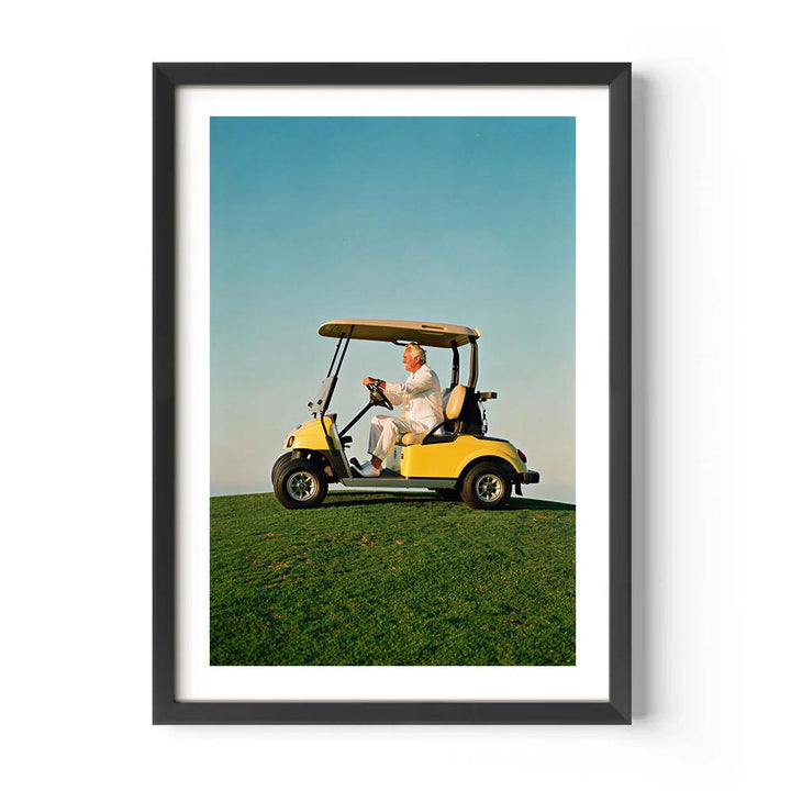 Framed artwork of a man driving a yellow golf cart on a green field with a blue sky.