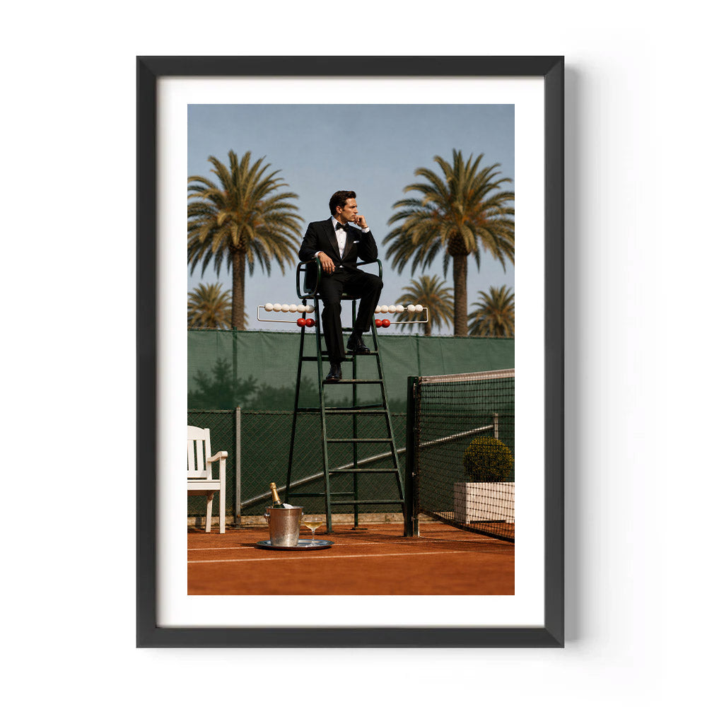 A framed art print featuring a man in a black tuxedo sitting on a tennis umpire chair on a clay tennis court, with palm trees and a tennis net in the background. The print is framed in black with a white mat, shown against a plain white wall.