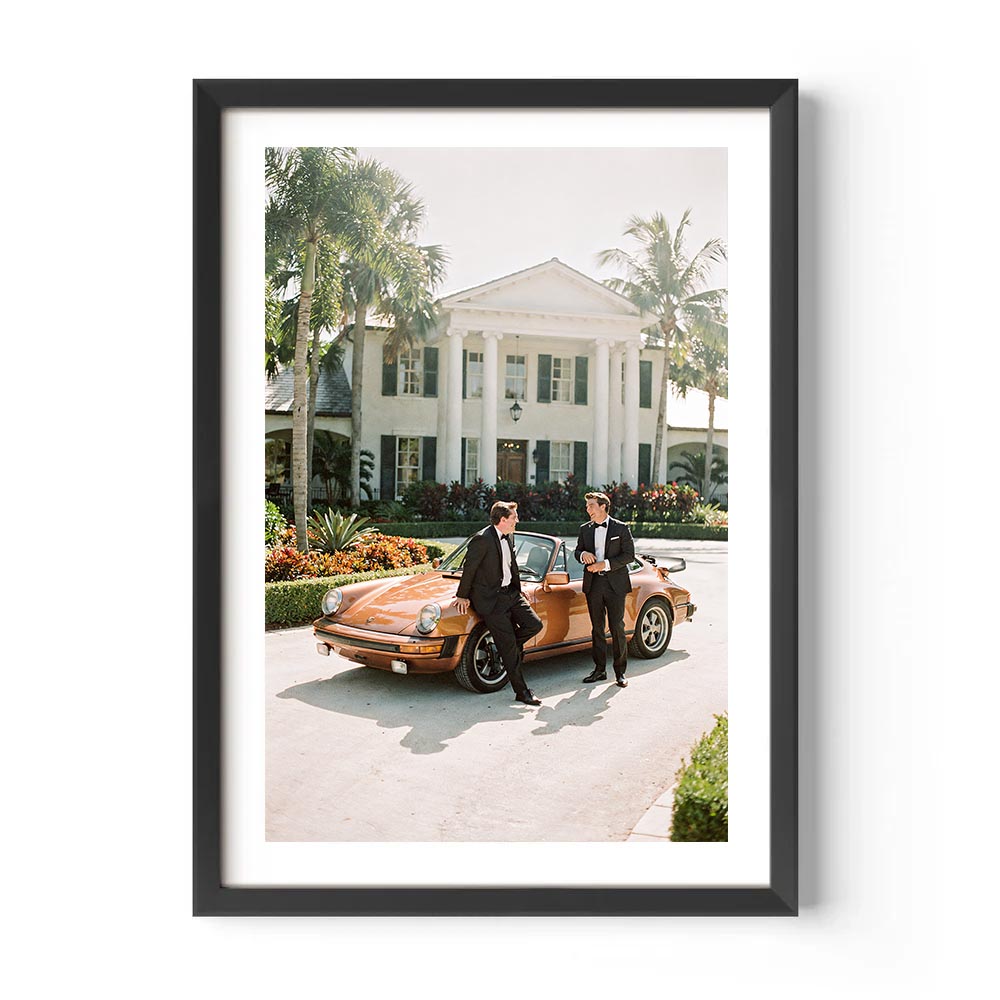 Two men in formal attire standing next to a vintage car in front of a large house with palm trees.