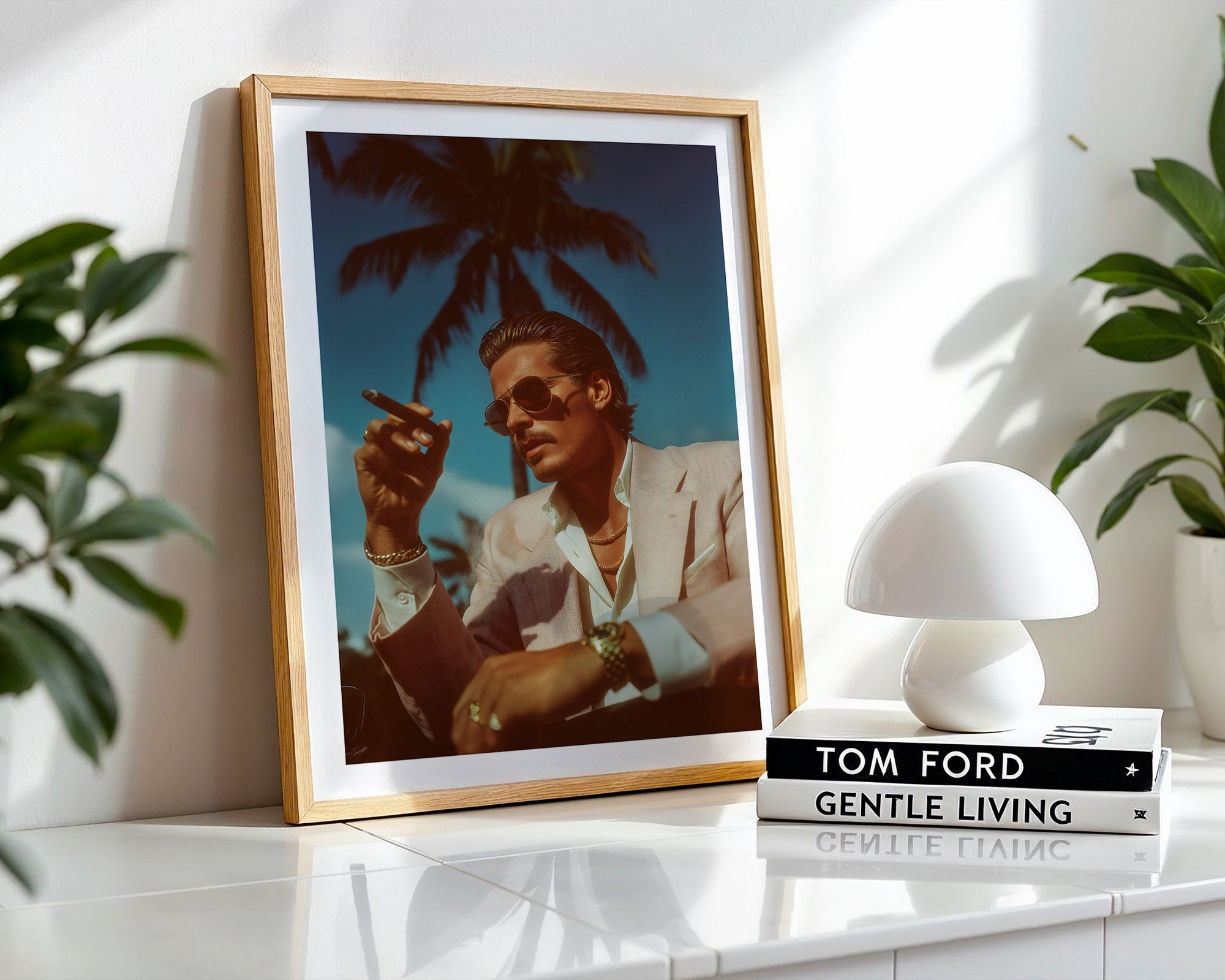 Framed portrait of a person with palm trees, placed on a surface with a white lamp and 'Tom Ford Gentle Living' book.