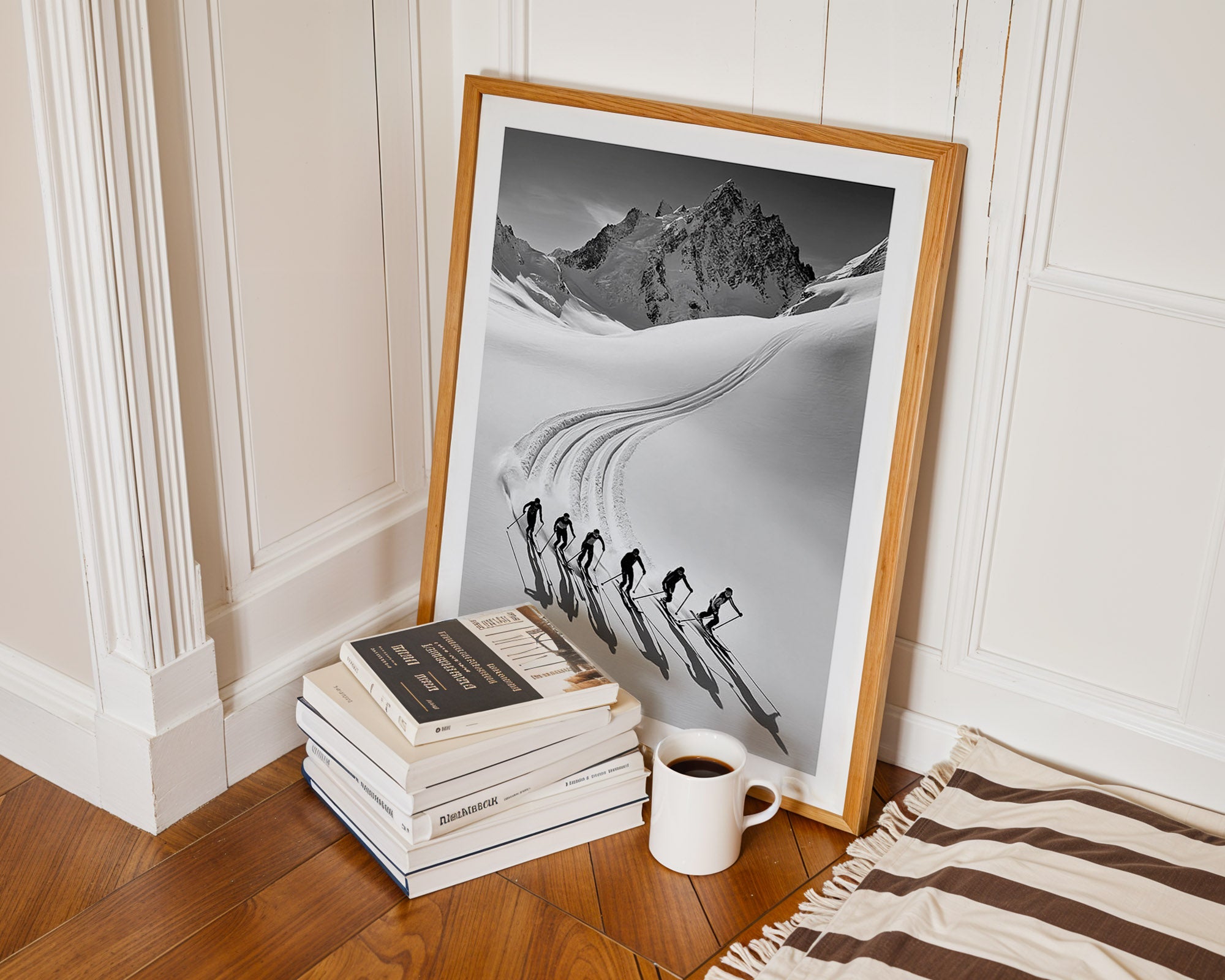 Framed black and white print of skiers on a mountain next to books and a mug on a wooden floor.