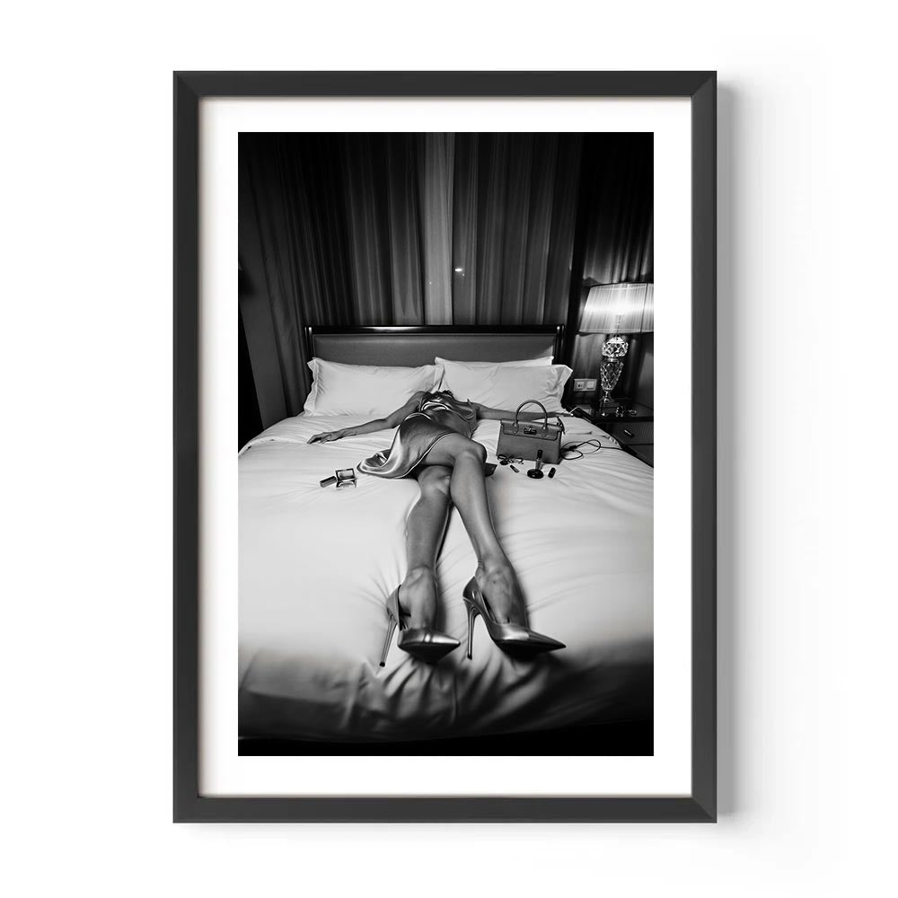 Framed black and white photo of a person lying on a bed with high heels and a purse.