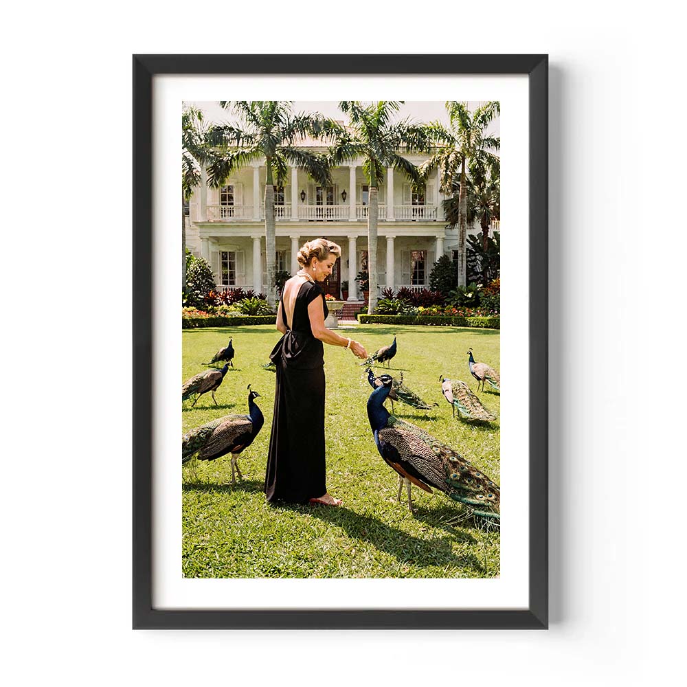 Framed photograph of a woman in a long black dress interacting with peacocks in front of a large house.