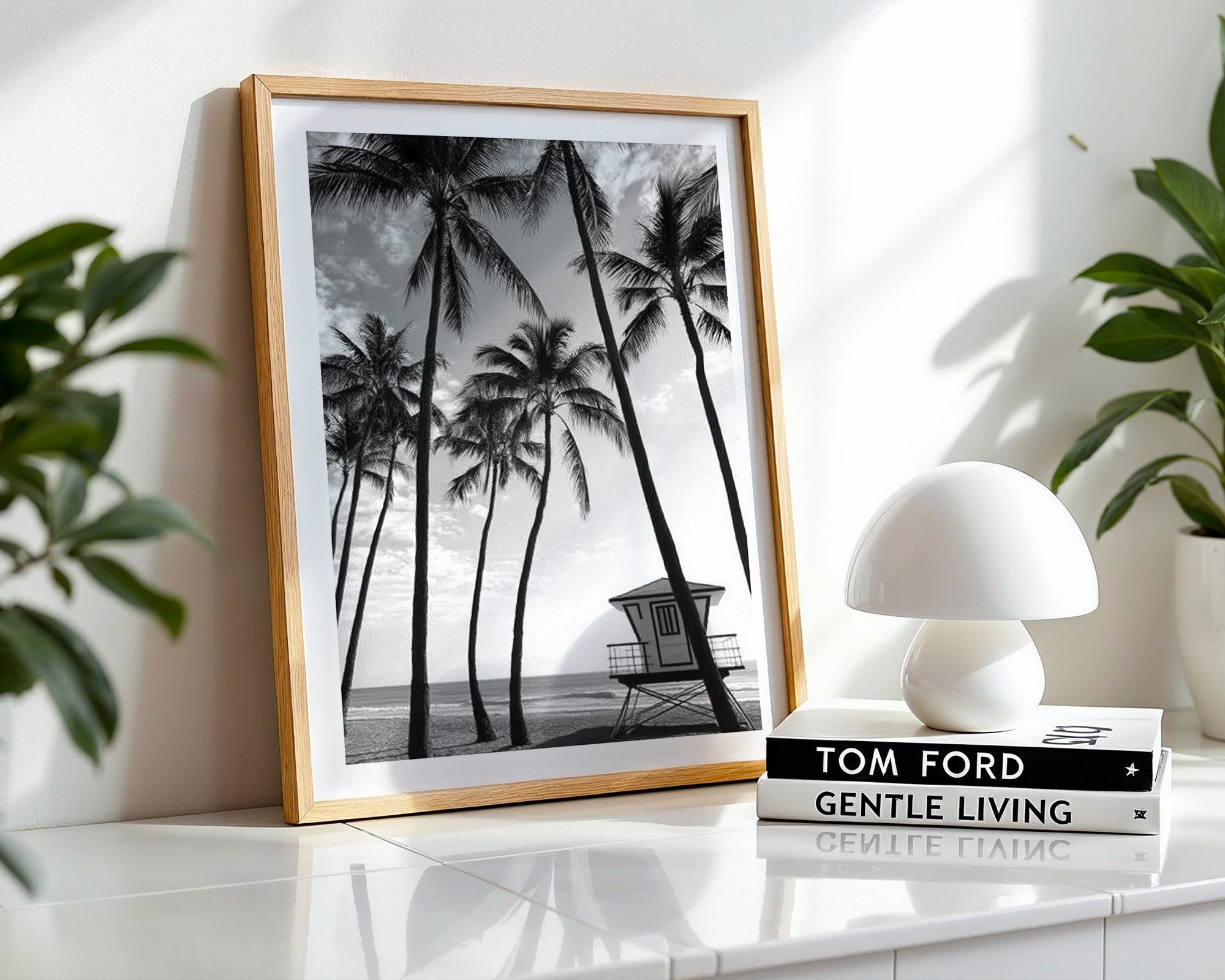 Framed black and white palm tree print on a shelf with books and a lamp.