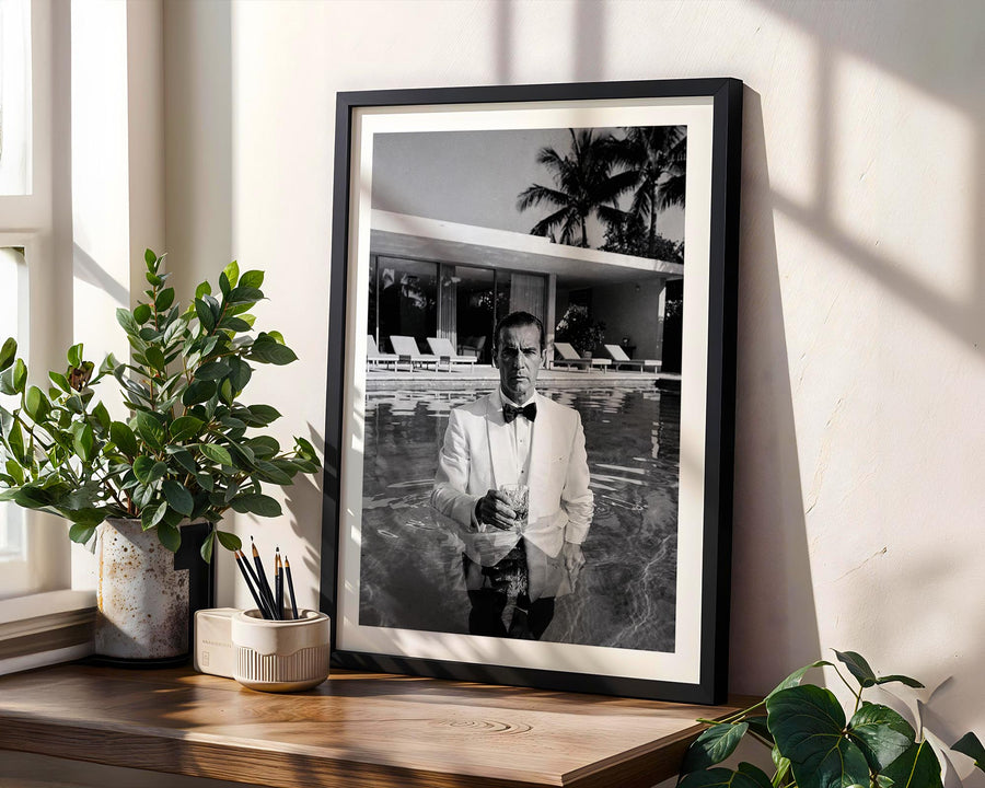 Framed black and white portrait of a man by a pool, placed on a wooden shelf with plants.
