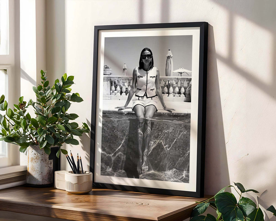 Framed black and white photograph of a woman sitting on a stone ledge, placed on a wooden shelf with plants.