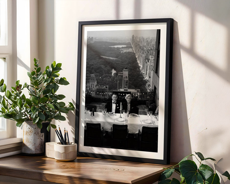 Framed black and white print of a cityscape on a wall with plants and a window.