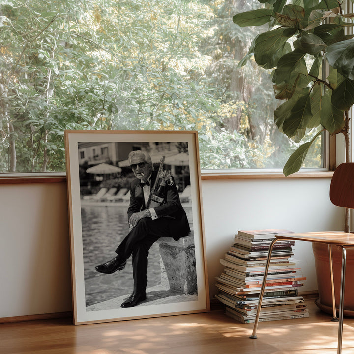 A framed black and white art print leaning against a window in a sunlit room, next to a wooden chair, a stack of books, and a large potted plant.