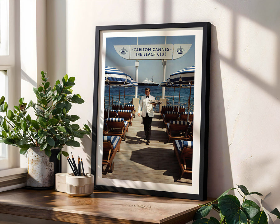 Framed poster of a beach club scene with a person walking on a wooden deck, placed on a windowsill with plants.