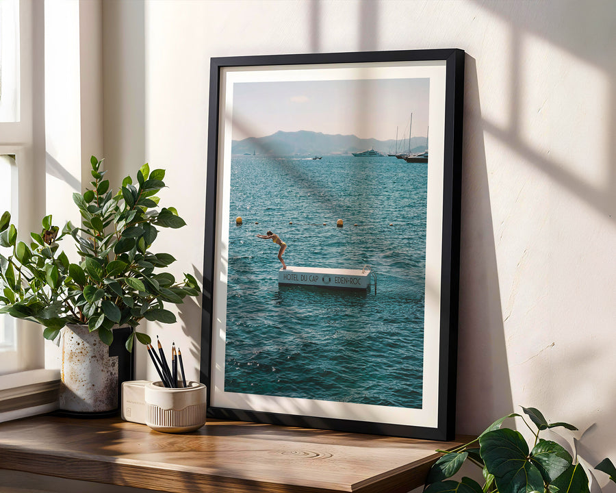 Framed photograph of a scenic view with a person on a boat, placed on a wooden surface with plants.
