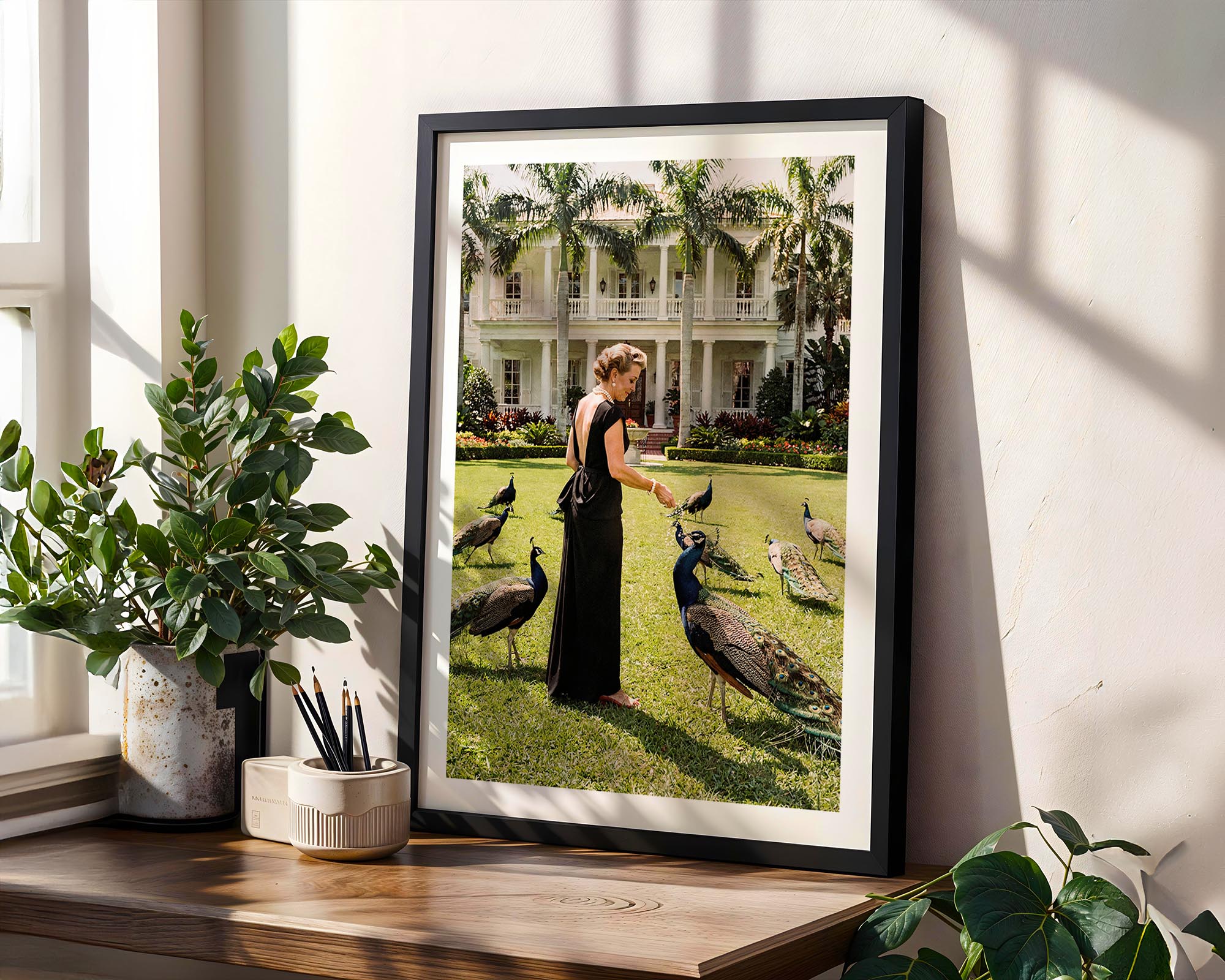 Framed photograph of a woman with peacocks in a garden setting on a wall next to plants.