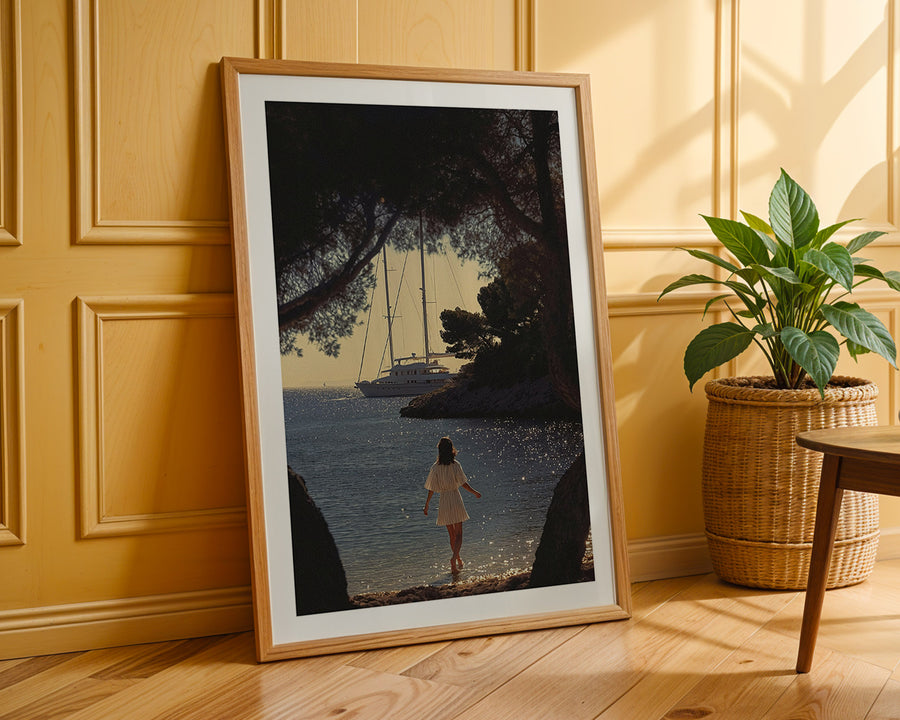 Framed artwork of a woman walking by a lake with a sailboat, placed on a wooden floor against a yellow paneled wall.