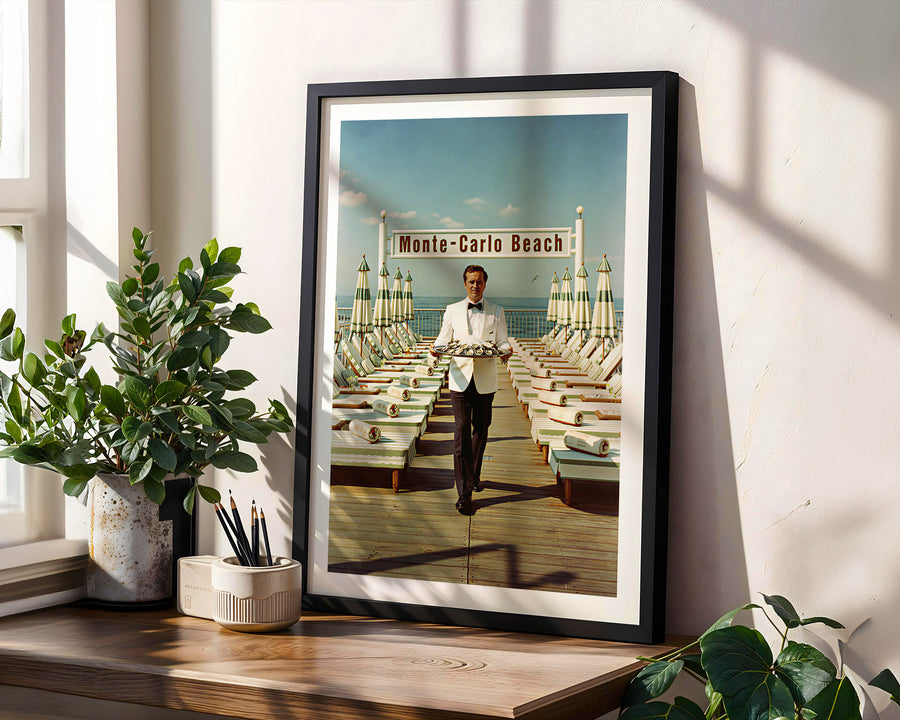 Framed artwork of a man on a dock with 'Monte-Carlo Beach' sign, placed on a wooden surface with plants.