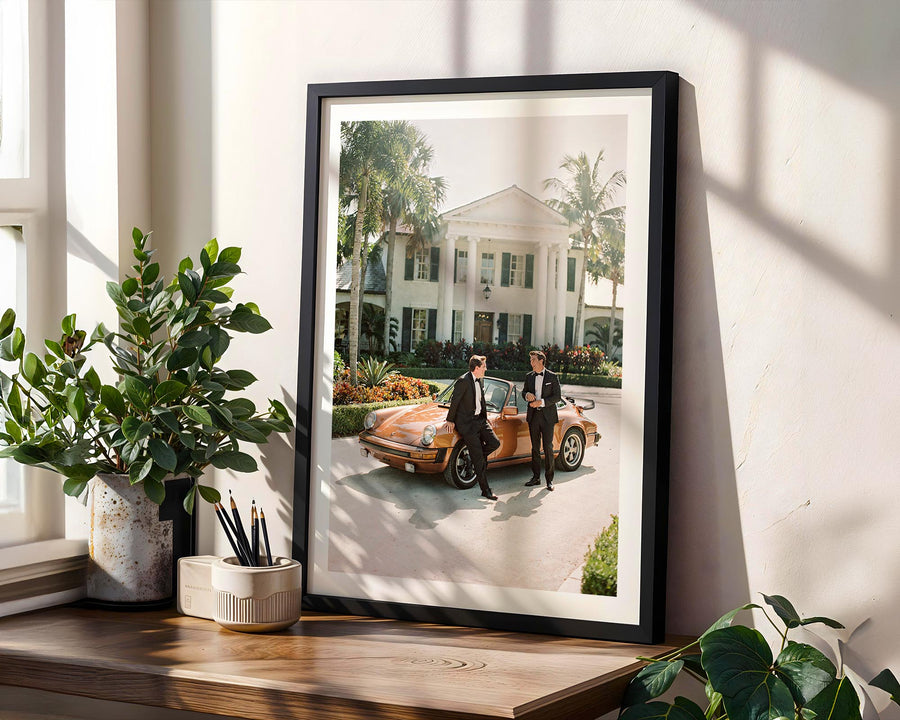 Framed photograph of two people by a vintage car on a wall with plants and a window in the background