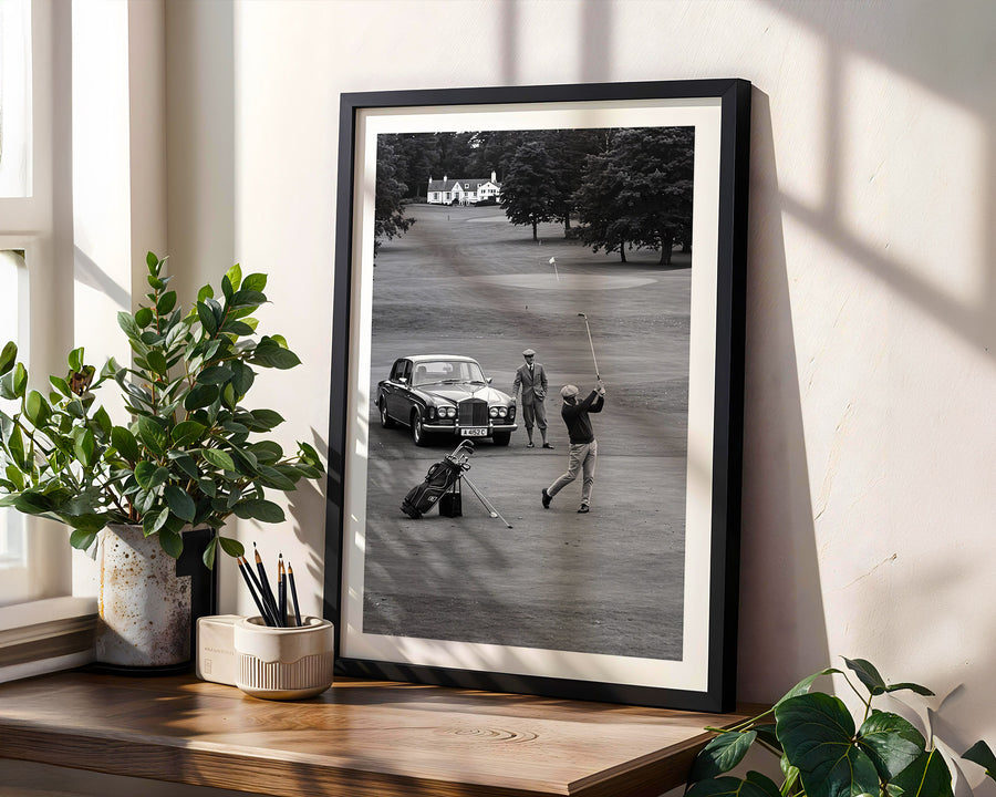Framed black and white photograph of a golfer on a golf course with a car in the background, displayed on a wooden shelf.