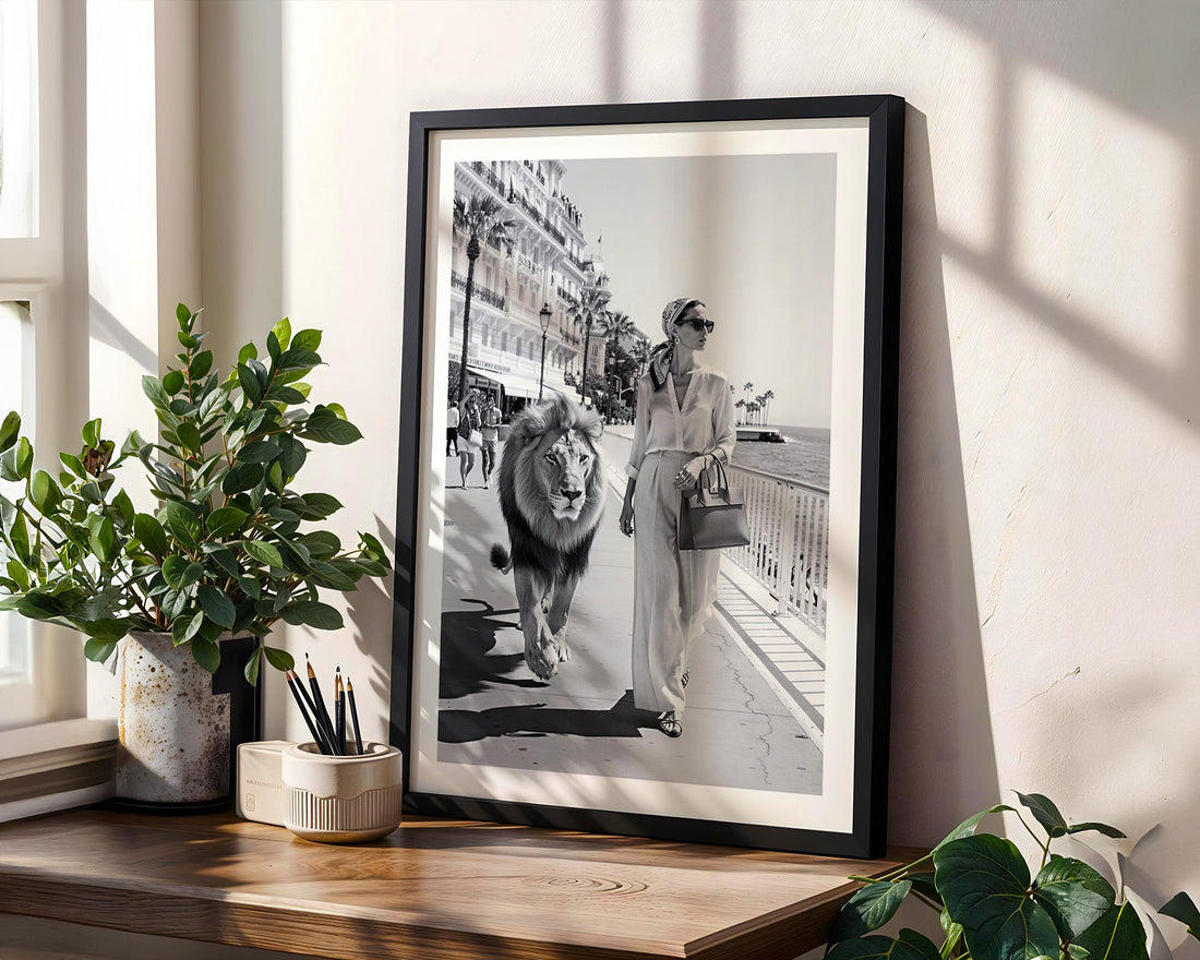 Framed black and white photo of a woman walking a lion on a city street, placed on a wooden surface with plants around.