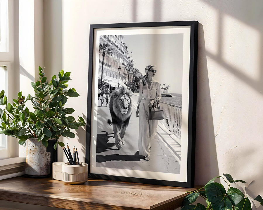 Framed black and white photo of a woman walking a lion on a city street, placed on a wooden surface with plants around.