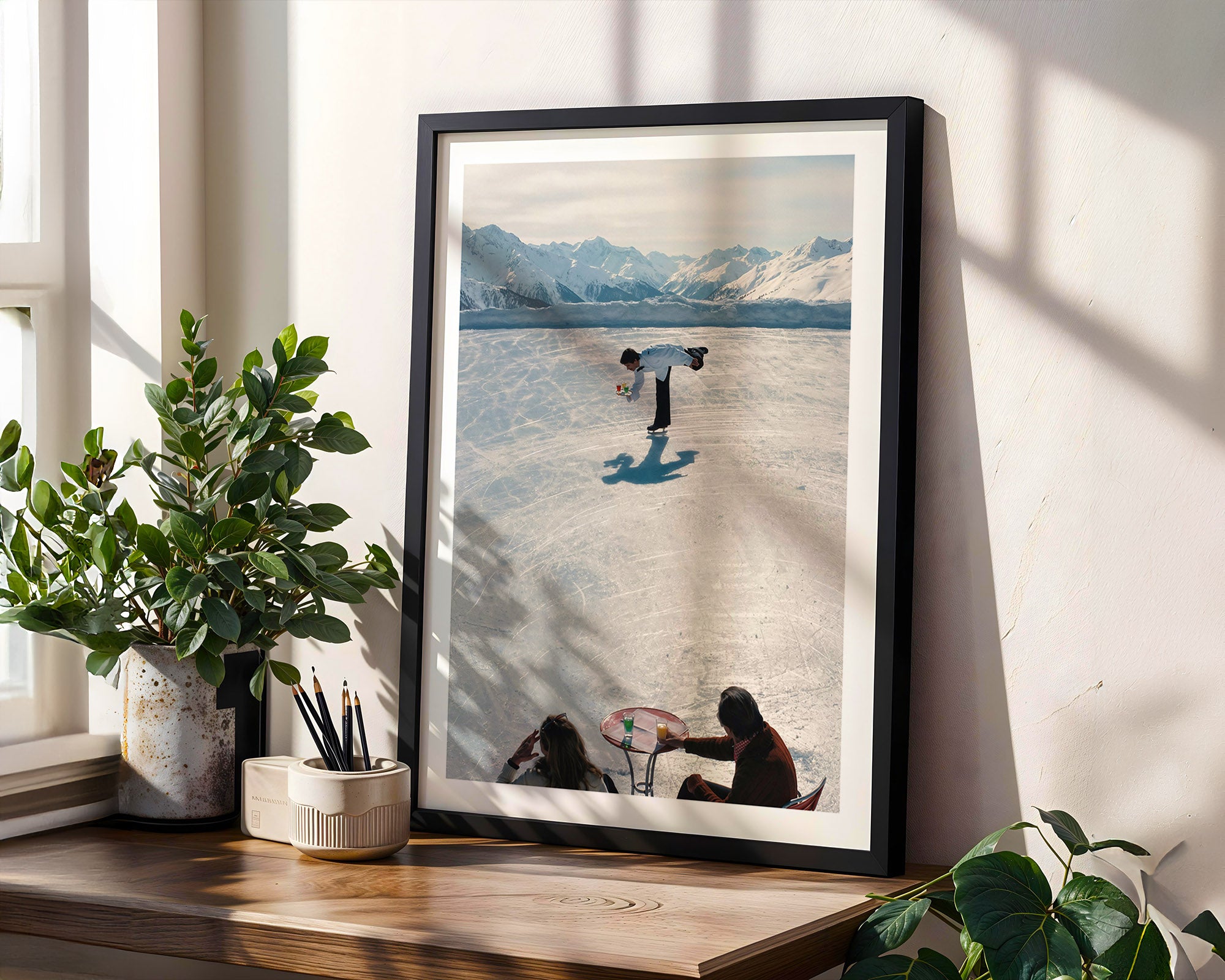 Framed artwork of a person skating on a lake with mountains in the background, displayed on a wooden shelf.