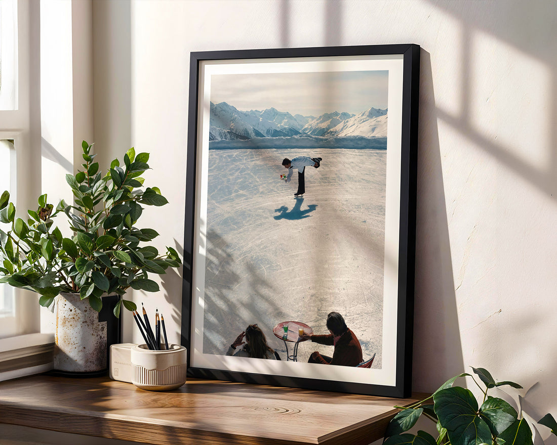 Framed artwork of a person skating on a lake with mountains in the background, displayed on a wooden shelf.