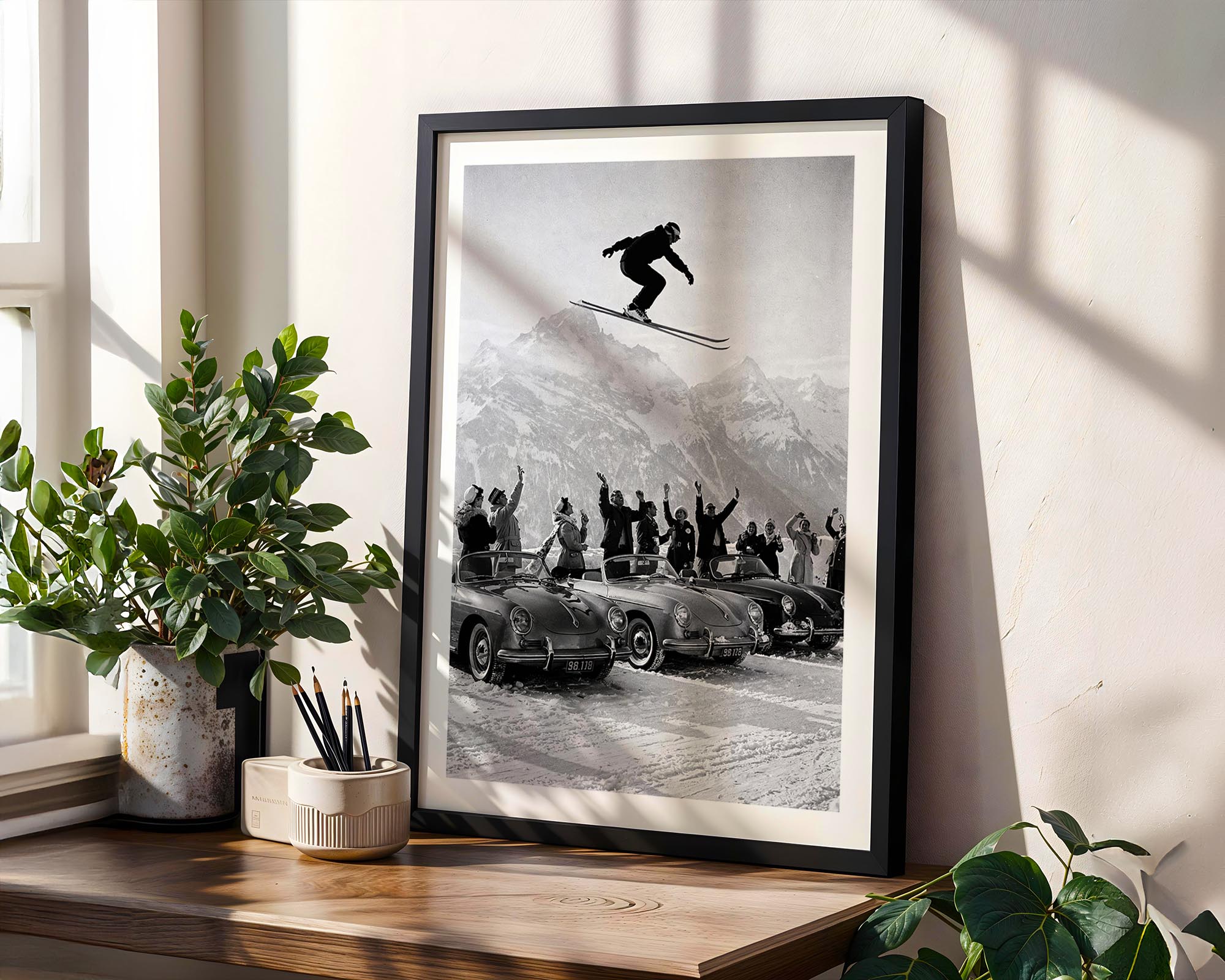 Framed black and white print of a skier in mid-air with a car and people below, on a wooden shelf with plants.