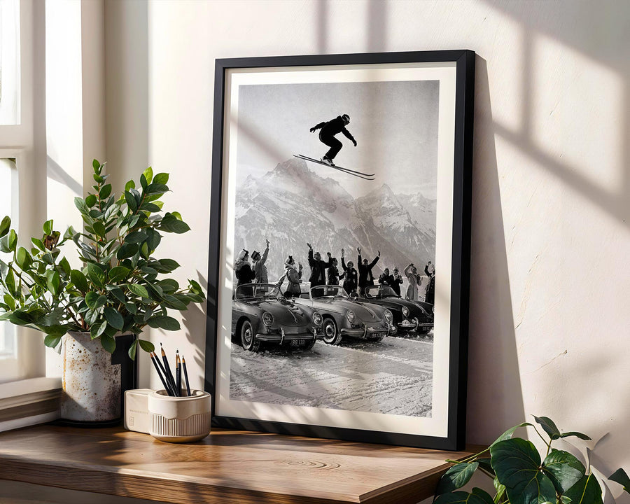 Framed black and white print of a skier in mid-air with a car and people below, on a wooden shelf with plants.
