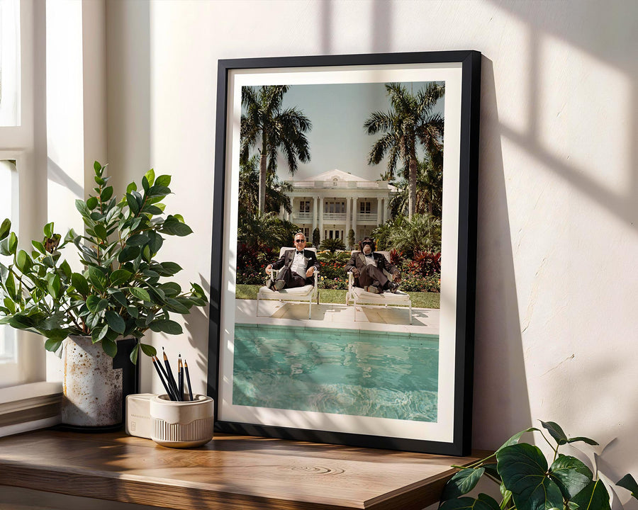 Framed photograph of a pool scene with people on a wooden shelf, surrounded by plants.