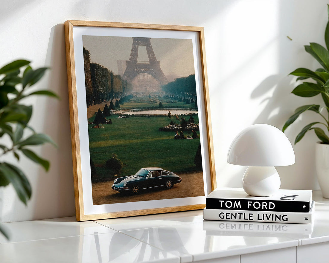 Framed artwork of a car and Eiffel Tower on a shelf with a white lamp and books.