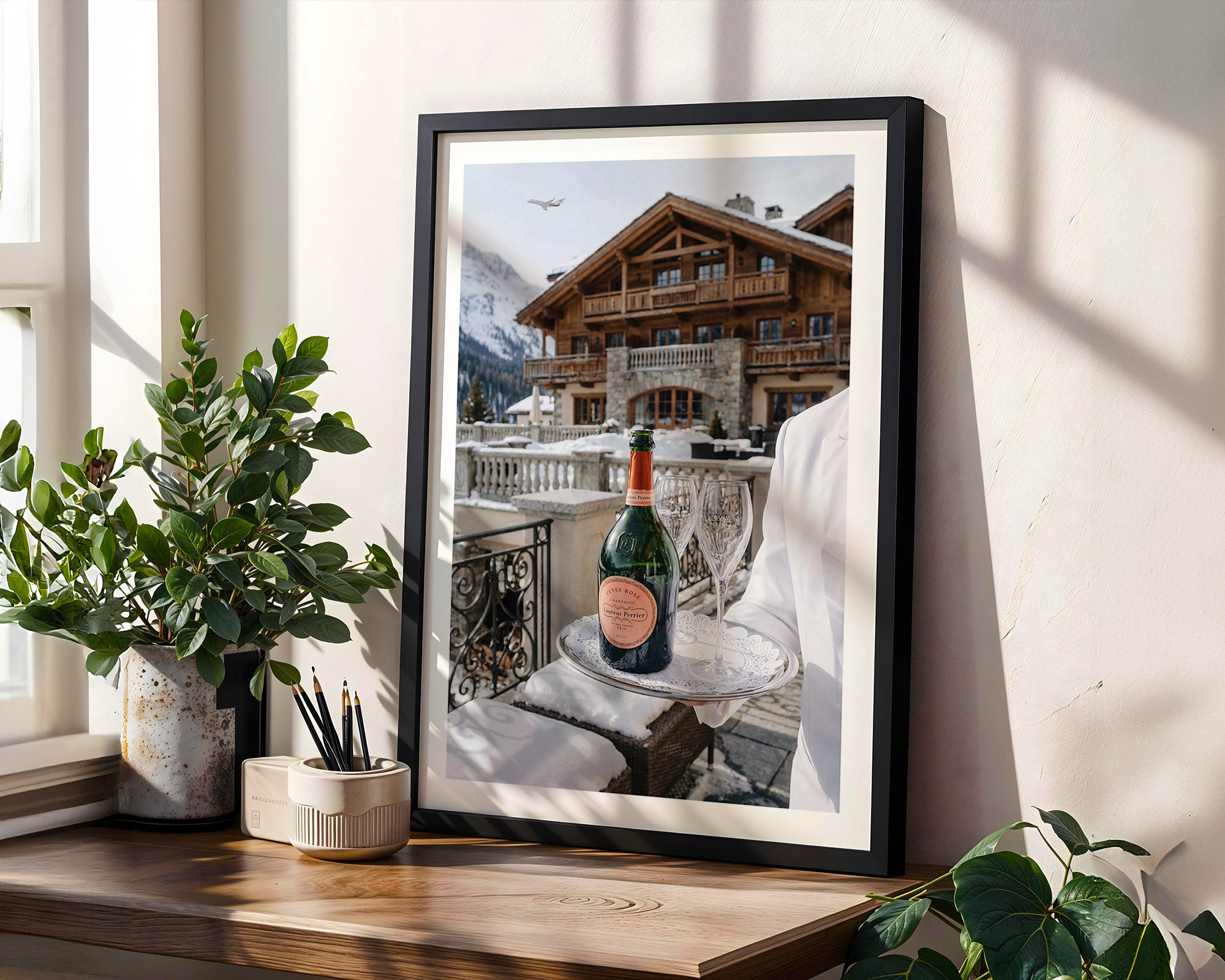 Framed photograph of a bottle on a table with a mountainous building in the background, placed on a wooden surface with plants.
