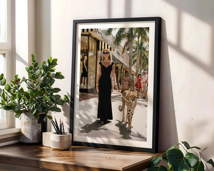 Framed photograph of a woman in a black dress walking a leopard on a city street, placed on a wooden surface with plants.