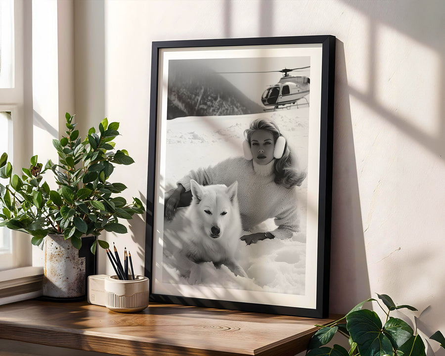 Framed black and white photo of a woman with a dog on a snowy landscape, placed on a wooden surface with plants.