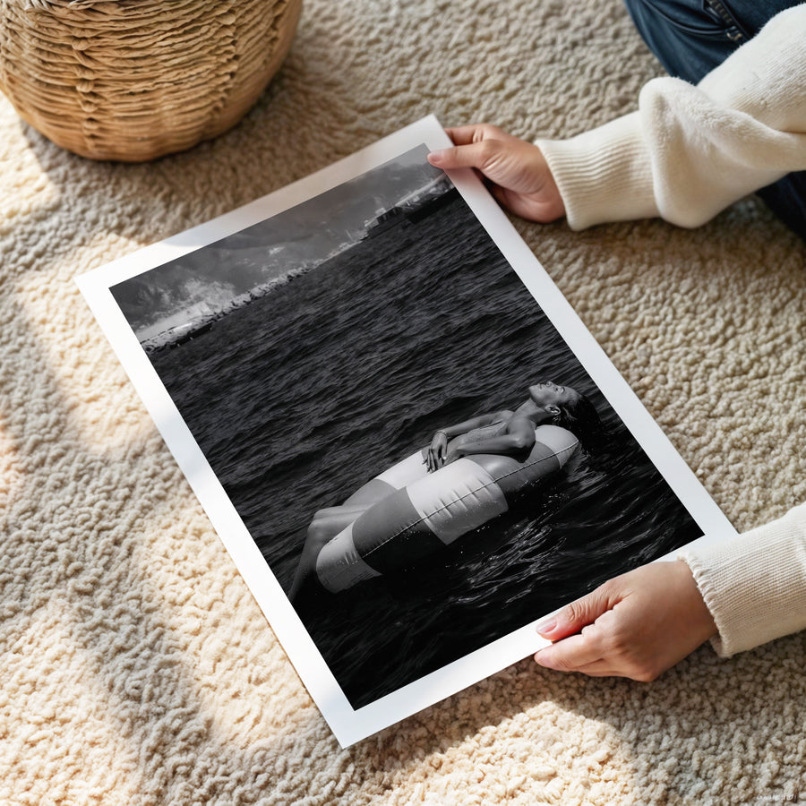 Person holding a framed black and white photograph of a person lying on a surfboard in the ocean.