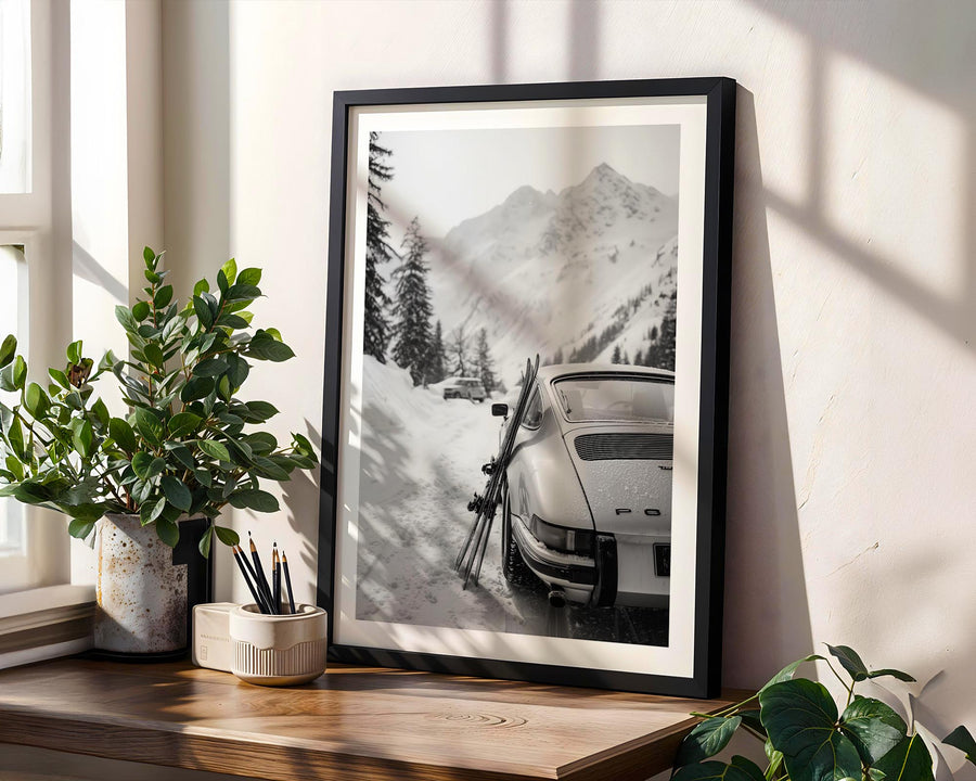 Framed black and white print of a snowy mountain scene with a Porsche 911 and skis, placed on a wooden surface with plants.