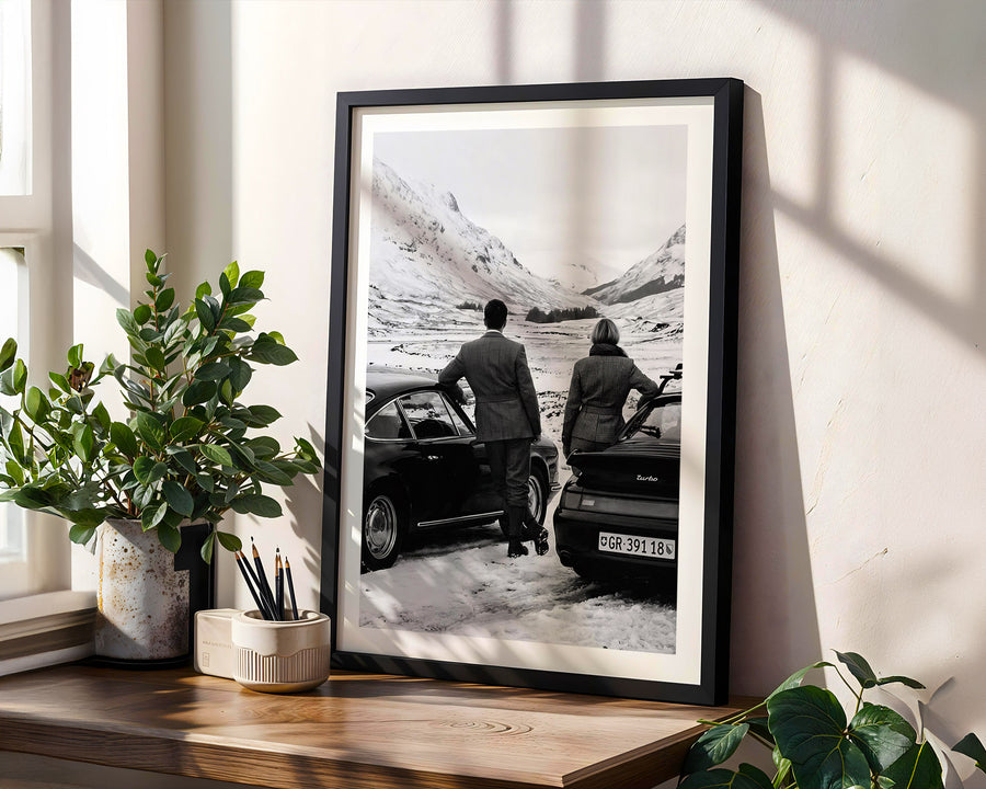 Framed black and white photograph of two people by a car on a mountain road, placed on a wooden shelf with plants.
