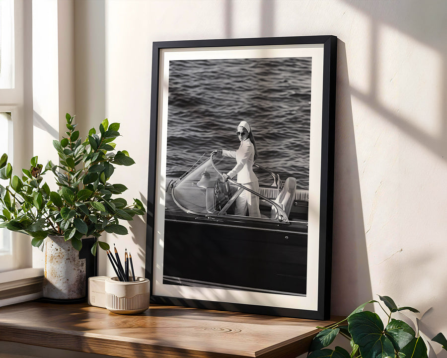 A framed black and white art print leaning against a wall on a wooden shelf, styled with potted plants and natural window light.