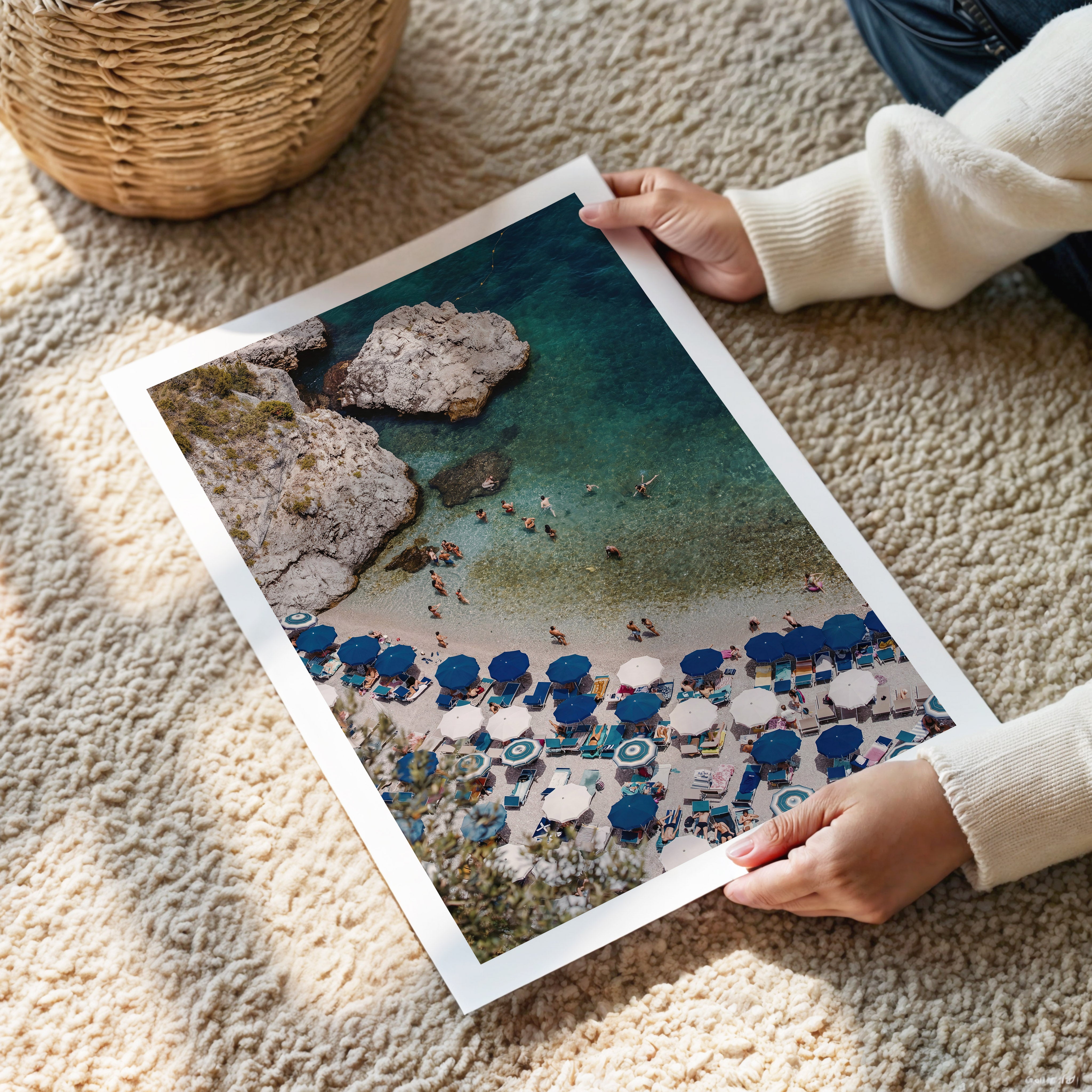 Person holding a large photo of a beach scene with blue umbrellas and rocky terrain.