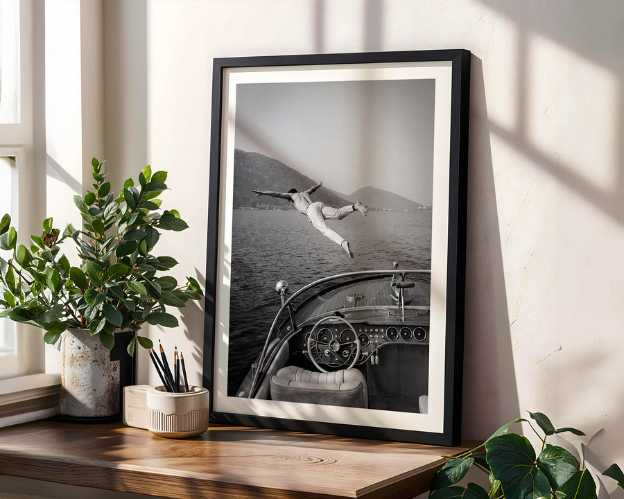 Framed black and white photograph of a person diving into water from a boat, placed on a wooden surface with plants.