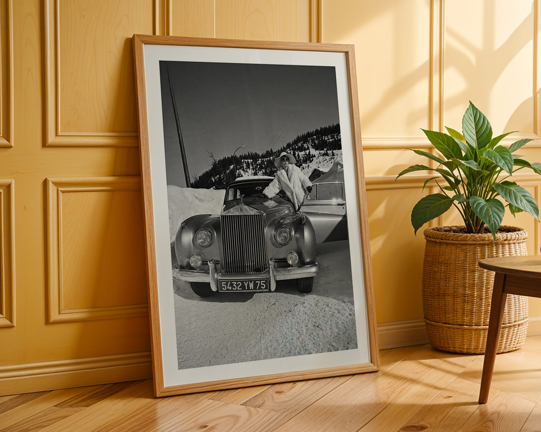Framed black and white photograph of a vintage car on a wooden floor with a plant in the background.
