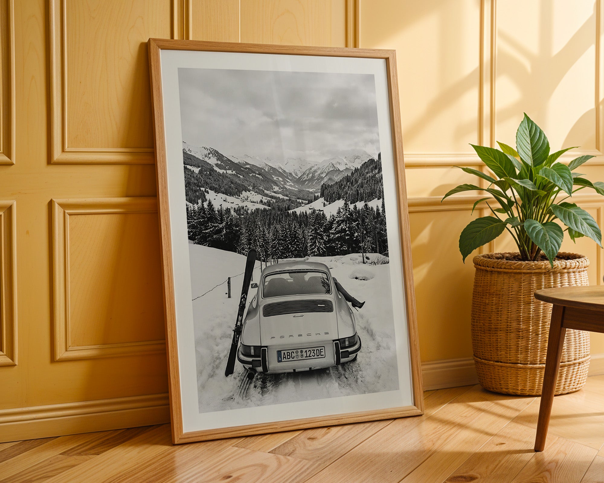 Framed black and white photograph of a snowy mountain scene with a car and skis, placed on a wooden floor against a yellow paneled wall.