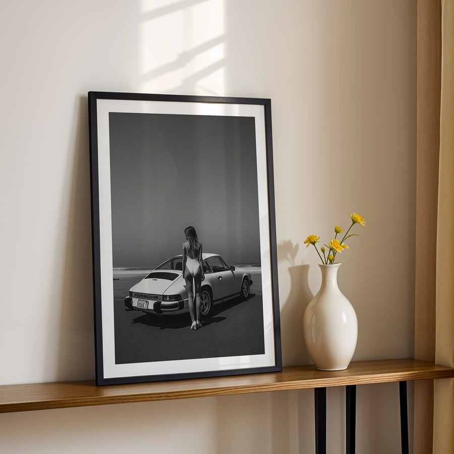Framed black and white photo of a woman by a car on a wooden shelf with a vase of flowers.