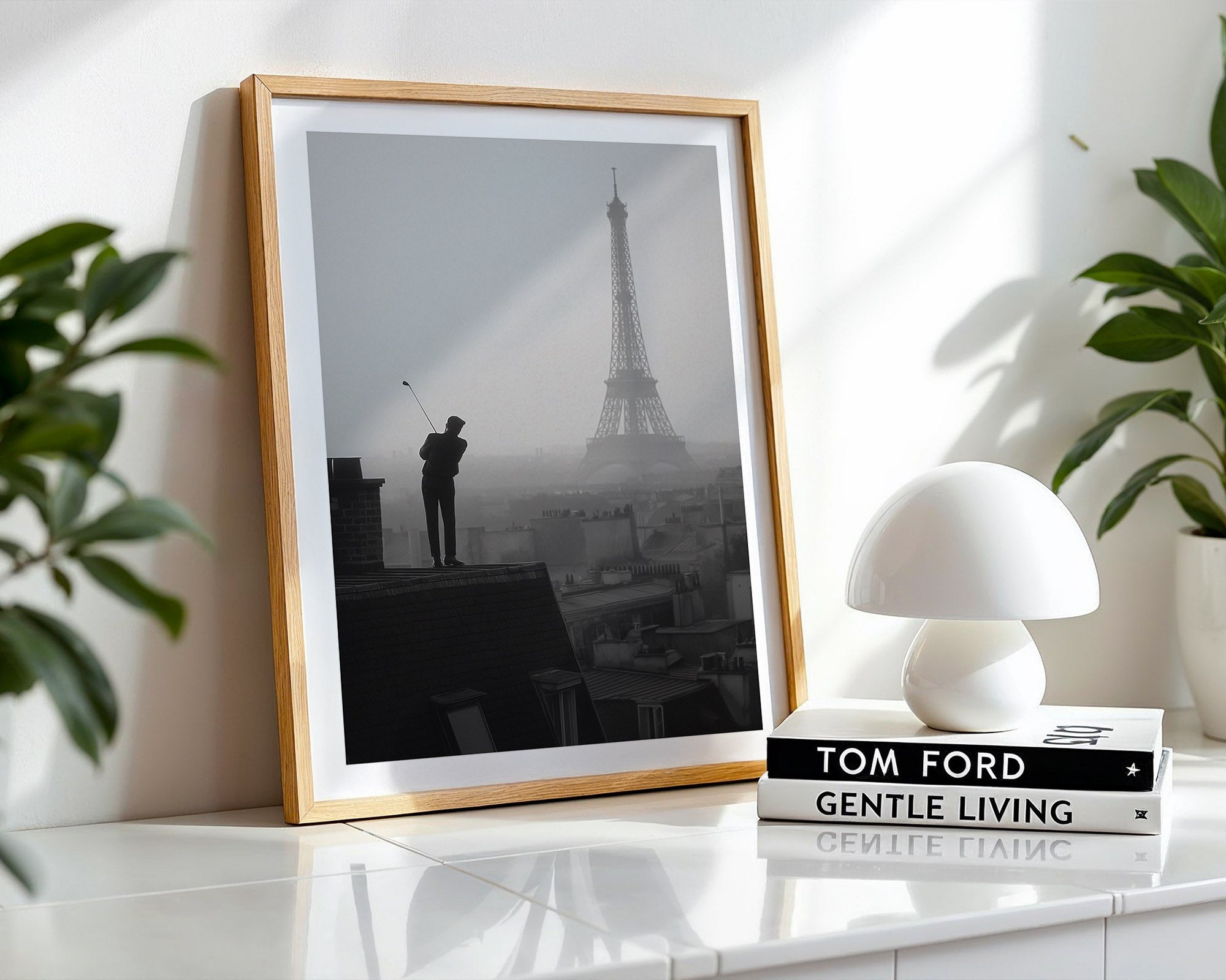 Framed artwork of a silhouette by the Eiffel Tower on a white surface with a lamp and books.