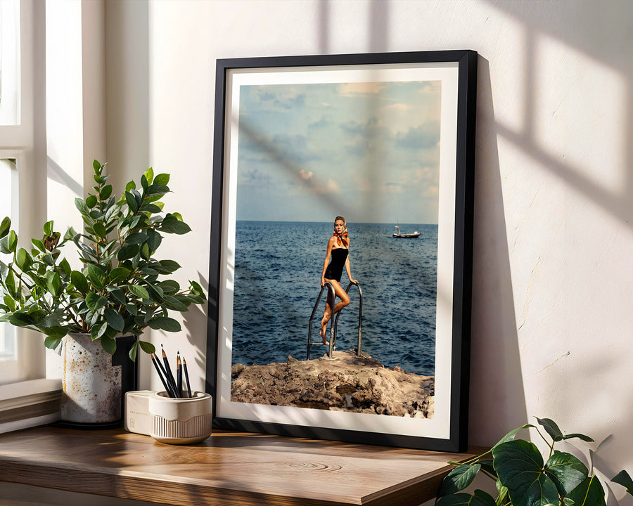 A framed art print leaning against a wall on a wooden shelf, next to potted green plants. The print features a vintage-style photograph of a woman in a black swimsuit standing on stilts on a rocky shoreline with the blue ocean and sky in the background.